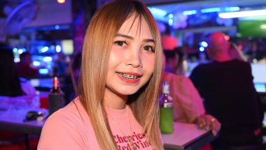 DZ6_0783: Young woman with long straight hair and braces smiling at a lively, neon-lit bar or club.