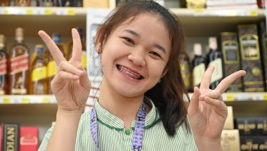 Media 'DZ6_0594: Smiling young woman flashes double peace signs while standing in a store aisle stocked with bottles on the shelves behind her.' in category 'Removable Appliances'