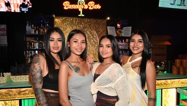 DZ6_2001: Four friends pose and smile together at the bar under the glowing "Bunny's Bar" sign, enjoying a night out.